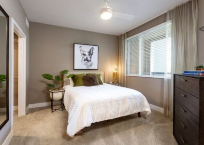 Anaheim CA Apartments for Rent - Rise - Cozy Bedroom with a Comfortable Bed, Carpet Flooring, a Ceiling Fan, and a Window with Blinds and Curtains